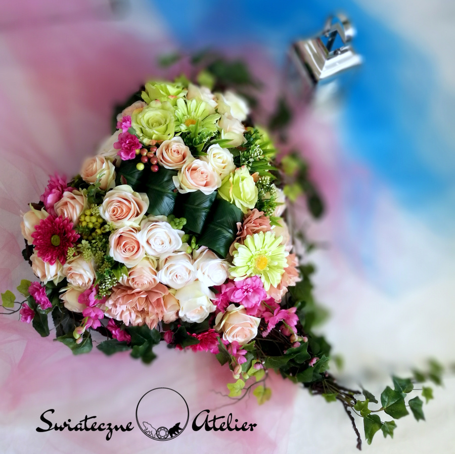 Nagrobne serce Czerwcowy dzień nr 763 Nagrobne serce Czerwcowy dzień nr 763 to wyjątkowa kompozycja na płytę nagrobną.  Kompozycja złożona z kilku kolorów róż i gerber, delikatnych listków i bluszczu tworzy harmonijną całość.  Główki gerber pięknie ożywiają kompozycję. Dekorację w całości wykonaliśmy ze sztucznych elementów, więc do złudzenia przypomina naturalną kompozycję.  Stroik nagrobny pięknie zdobią zielone sztuczne gałązki. Ze względu na kolorystykę oraz wykorzystane materiały, znakomicie zaprezentuje się na nagrobku. Serce jest duże i okazałe o wiosennej kolorystyce.